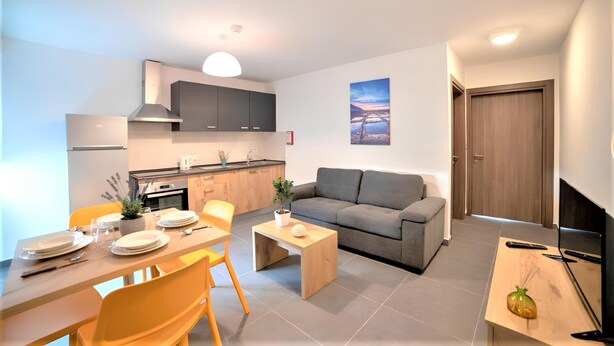 Una sala moderna con un sofá, una mesa de comedor y sillas. Hay una zona de cocina con armarios y una estufa. Un televisor está sobre un mueble bajo, y una planta está sobre la mesa.