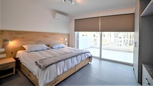 Un dormitorio moderno con cabecera de madera, una cama grande con ropa blanca, una mesita de noche, aire acondicionado de pared y un ventanal con vistas a edificios.