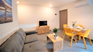 Una sala moderna con un sofá gris, una mesa de centro de madera y un televisor sobre un mueble de madera. Hay un comedor con sillas amarillas y una mesa preparada para dos personas.