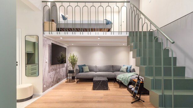 Ein modernes Wohnzimmer mit einem grauen Sofa, einem kleinen Tisch, einer Treppe und einem Balkon mit Geländer.