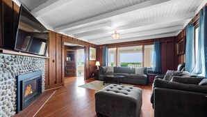 Ein Wohnzimmer mit Kamin, einer Sofagarnitur, einem Fernseher und einem großen Fenster mit Vorhängen.