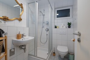 Un baño moderno con ducha envuelta en vidrio, un lavamanos blanco, inodoro y espejo con marco decorativo.