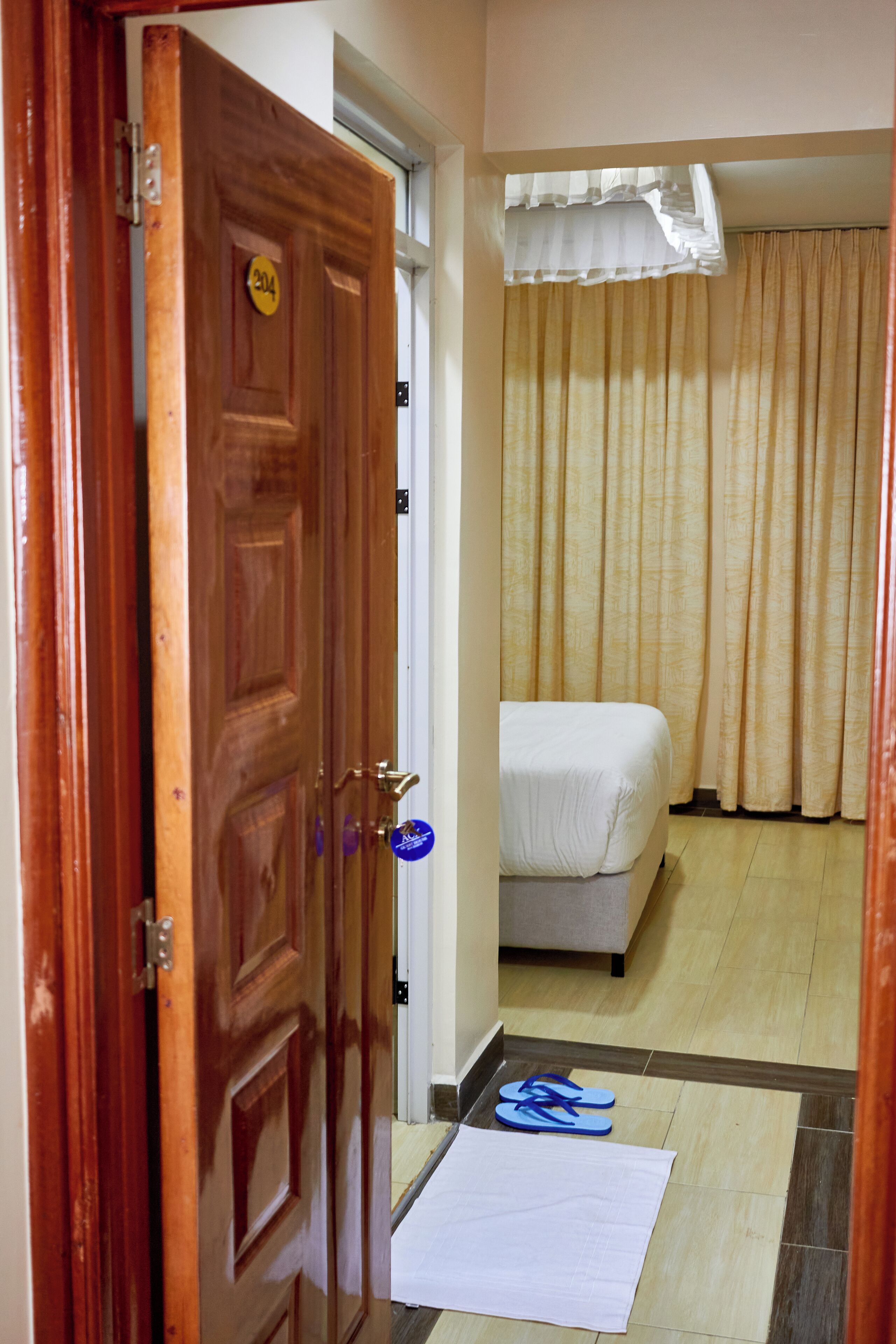 Ein Hotelzimmer mit Holztür, einem Bett, einem Vorhang und einem Paar Hausschuhen auf dem Boden.