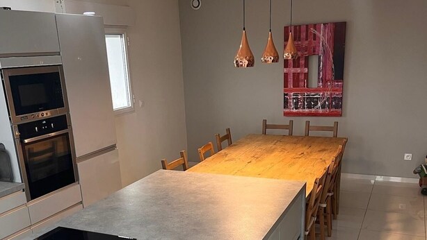 A modern kitchen with a large island, built-in ovens, pendant lights, and a dining area with a long table. - Modern apartment with incredible views (Le Crès)
