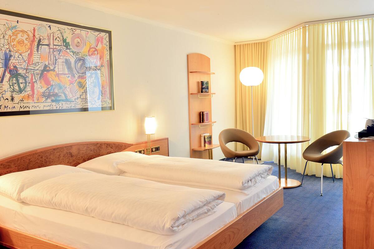 Camera d'albergo con un letto matrimoniale, una scrivania con due sedie, una libreria e un grande quadro astratto appeso alla parete.