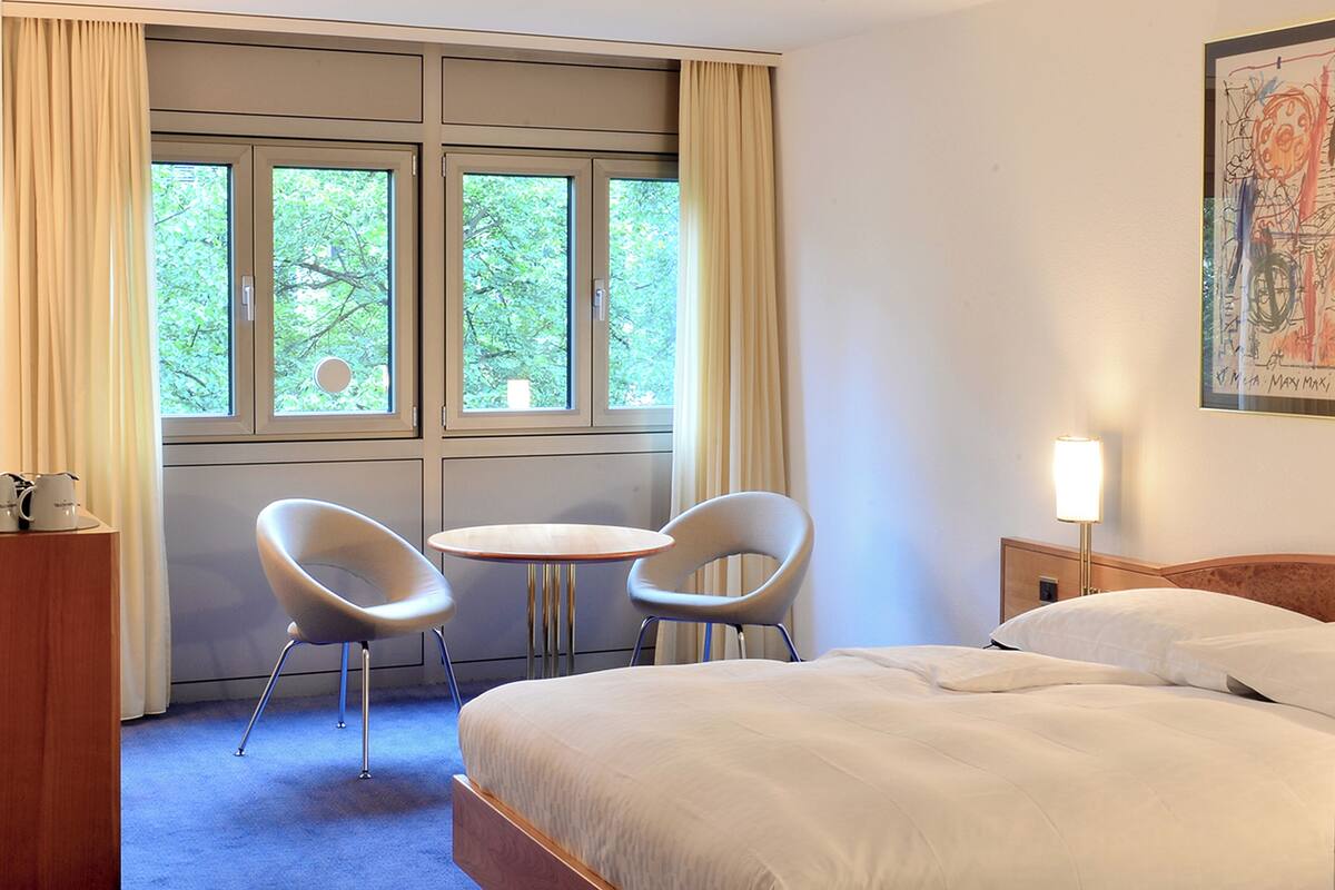 Una camera d'albergo con un letto, due sedie, un tavolino, una lampada e una finestra con tende.