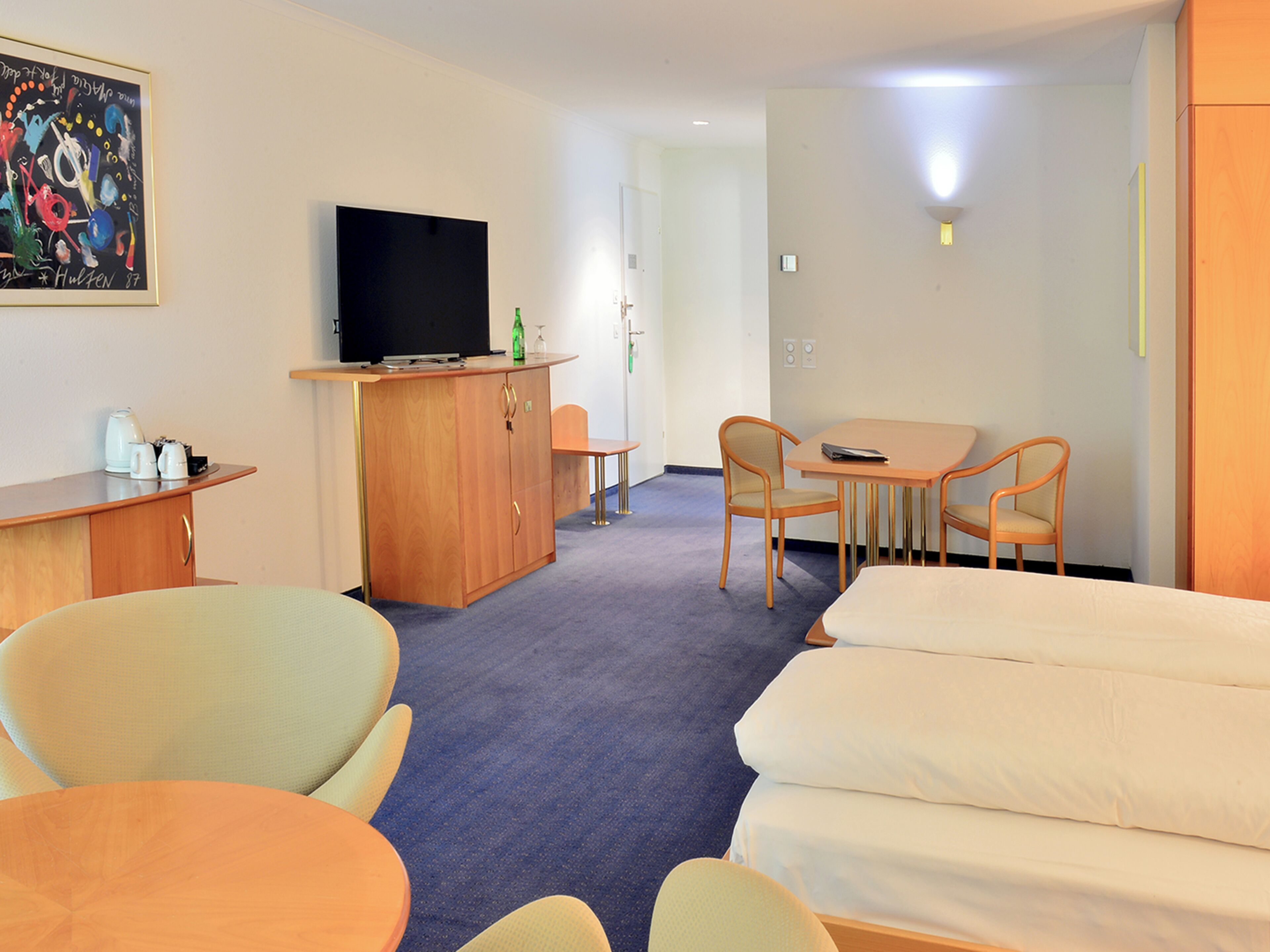 A hotel room with a bed, a desk with a television, a dining area with chairs, and a painting on the wall.