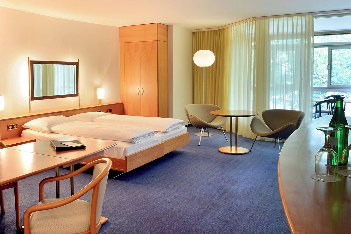 Camera d'albergo con un letto, due sedie, un tavolino, una scrivania, una lampada e una bottiglia sul tavolo.