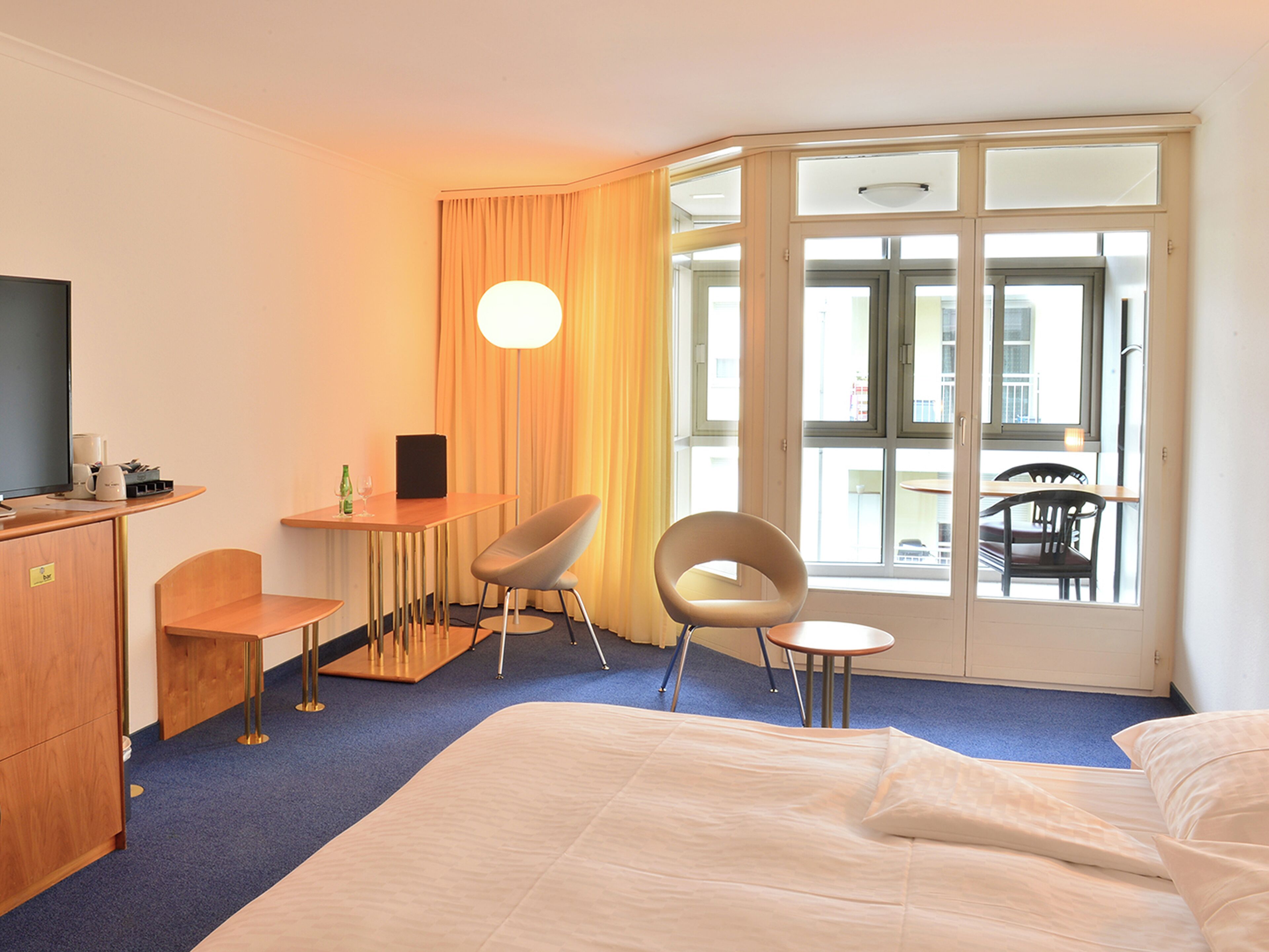 A hotel room with a bed, two chairs, a desk, a television, and a large window with curtains.