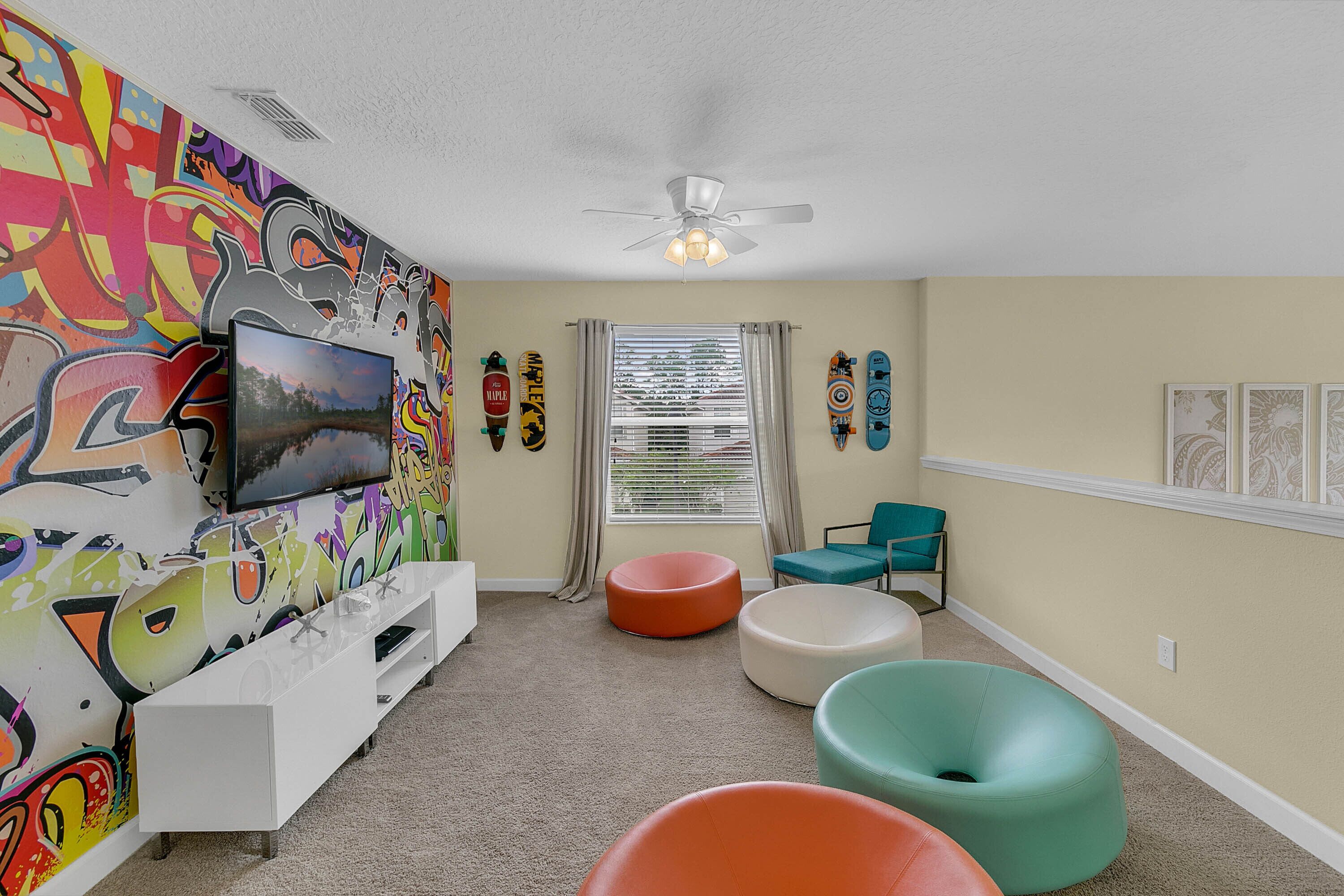 Un salon avec un mur décoré de graffitis colorés, un téléviseur à écran plat, un ventilateur de plafond et une fenêtre avec des stores.