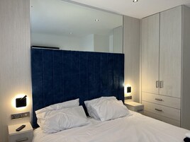 A modern hotel room with a large bed, a nightstand with a remote control, a wardrobe with a mirror, and wall-mounted lighting.