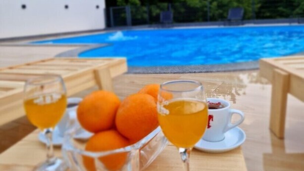 Een zwembadterras met een houten tafel, een glas sinaasappelsap, een glas thee en een schaal mandarijnen.