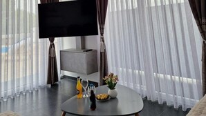 Een moderne woonkamer met een flatscreen televisie, een grijze bank, een ronde salontafel met drankjes en snacks, en grote ramen met dunne gordijnen.