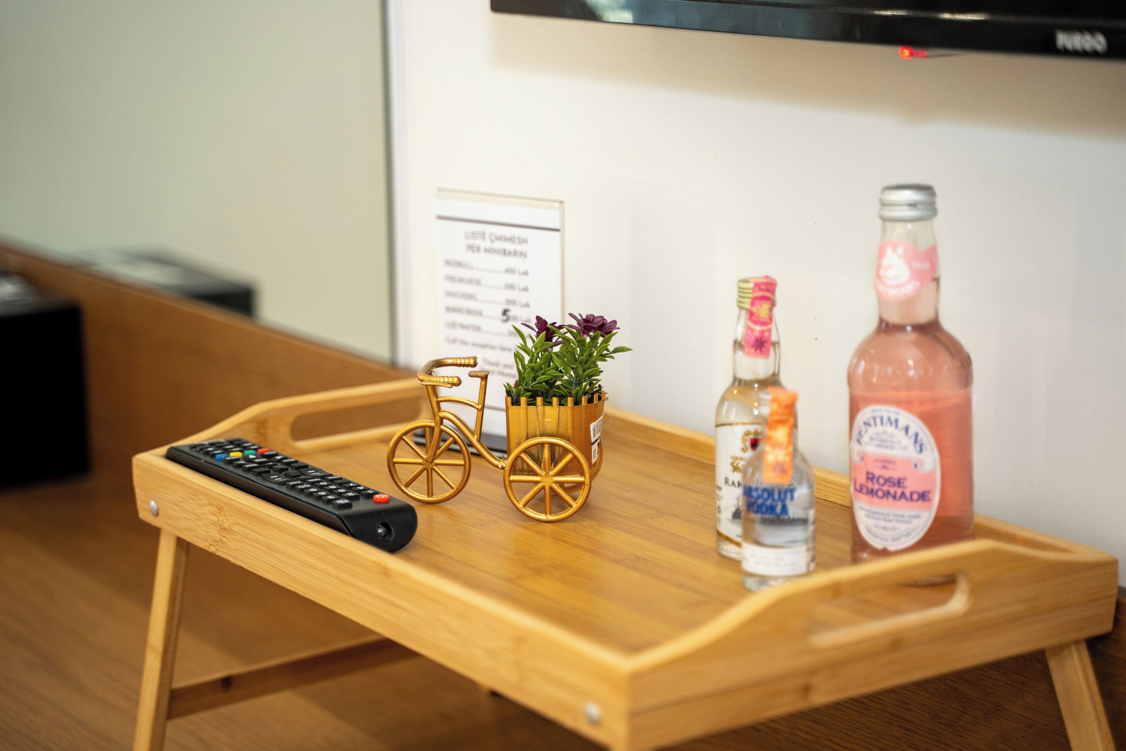Uma bandeja de madeira com um controle remoto, um vaso em formato de bicicleta, uma garrafa de vodka e uma garrafa de limonada de rosas.