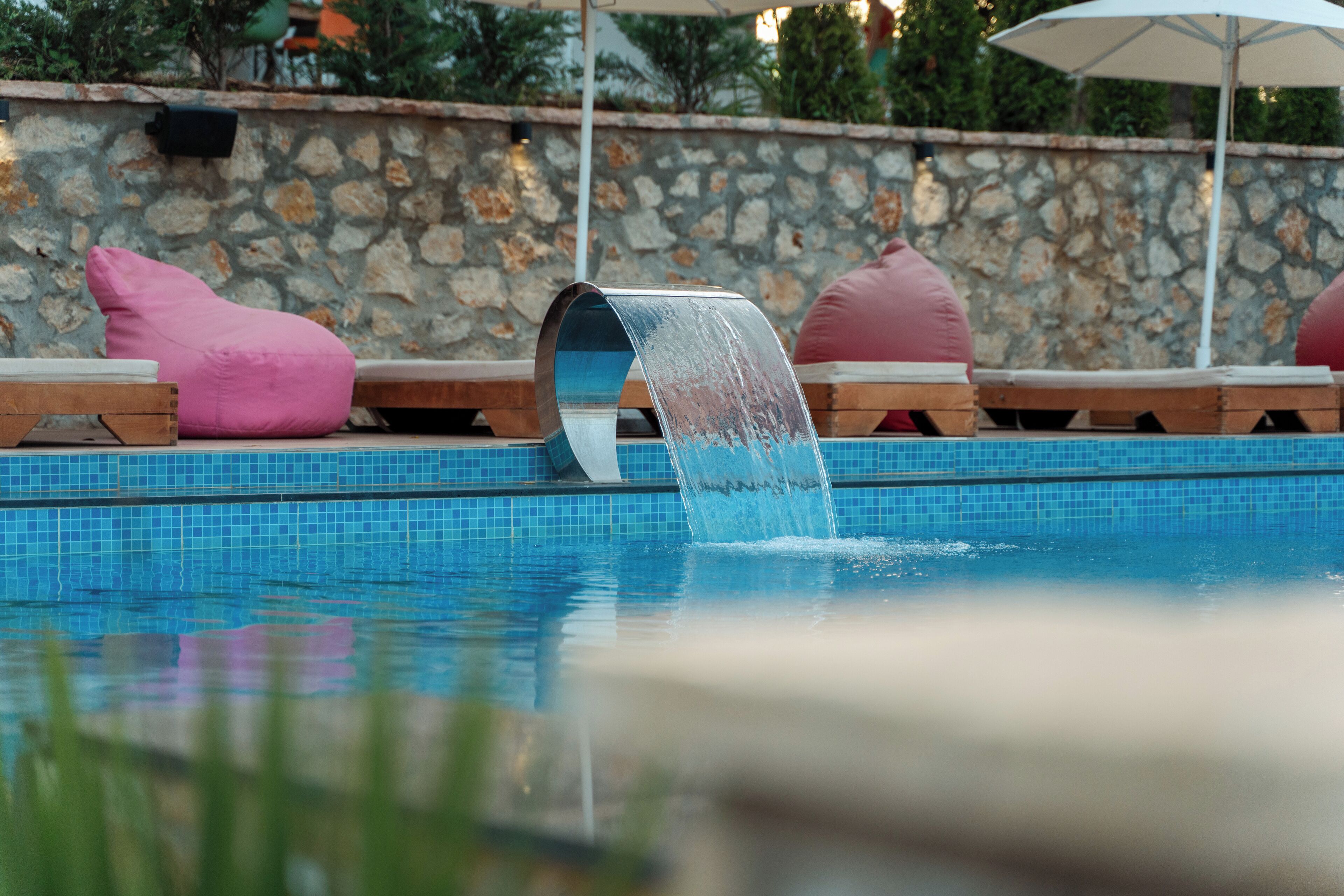 Una piscina con elemento d'acqua, parete in pietra e cuscini rosa sulle poltrone prendisole.