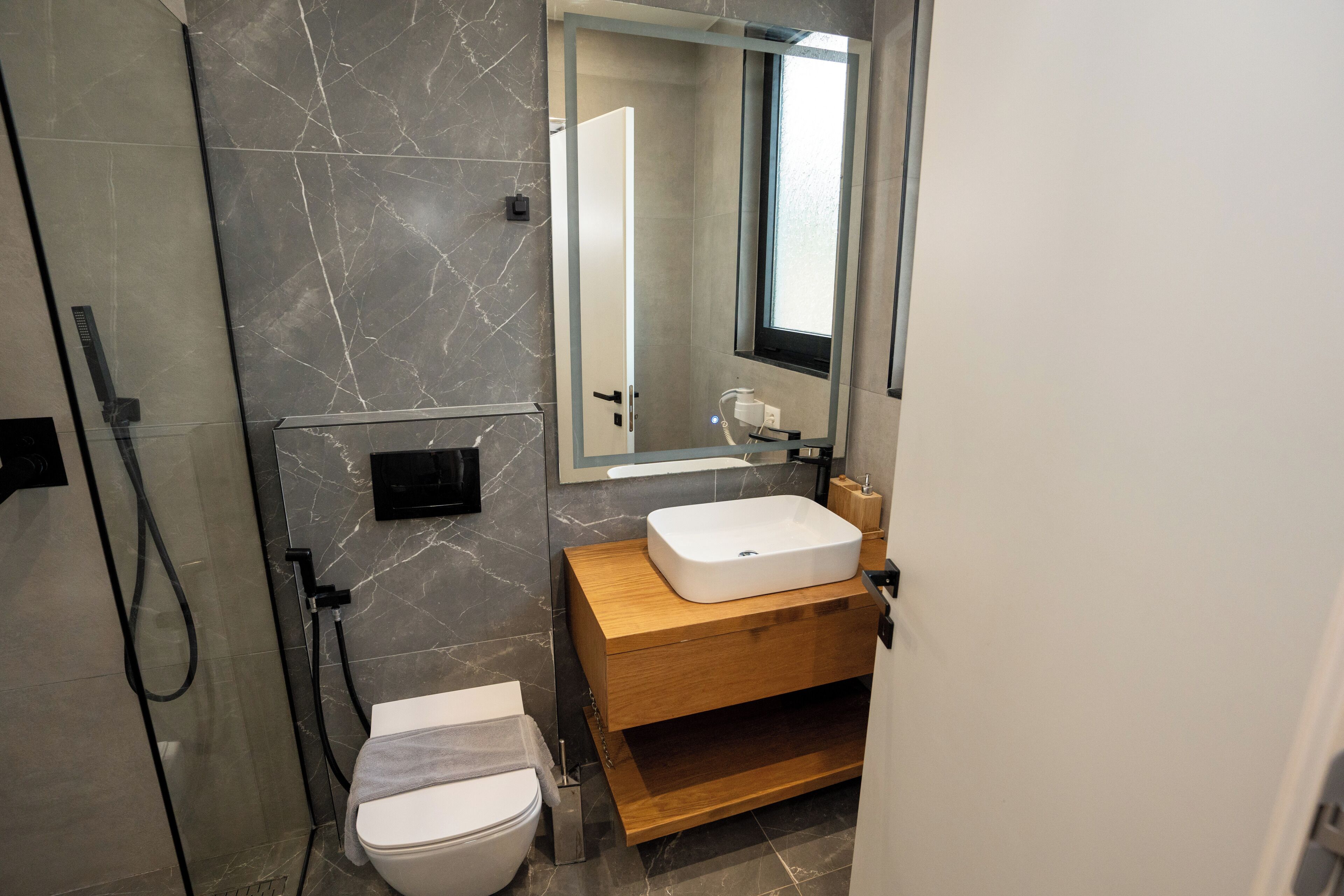 A modern bathroom with a marble wall, a wooden vanity, a white sink, and a glass shower enclosure.