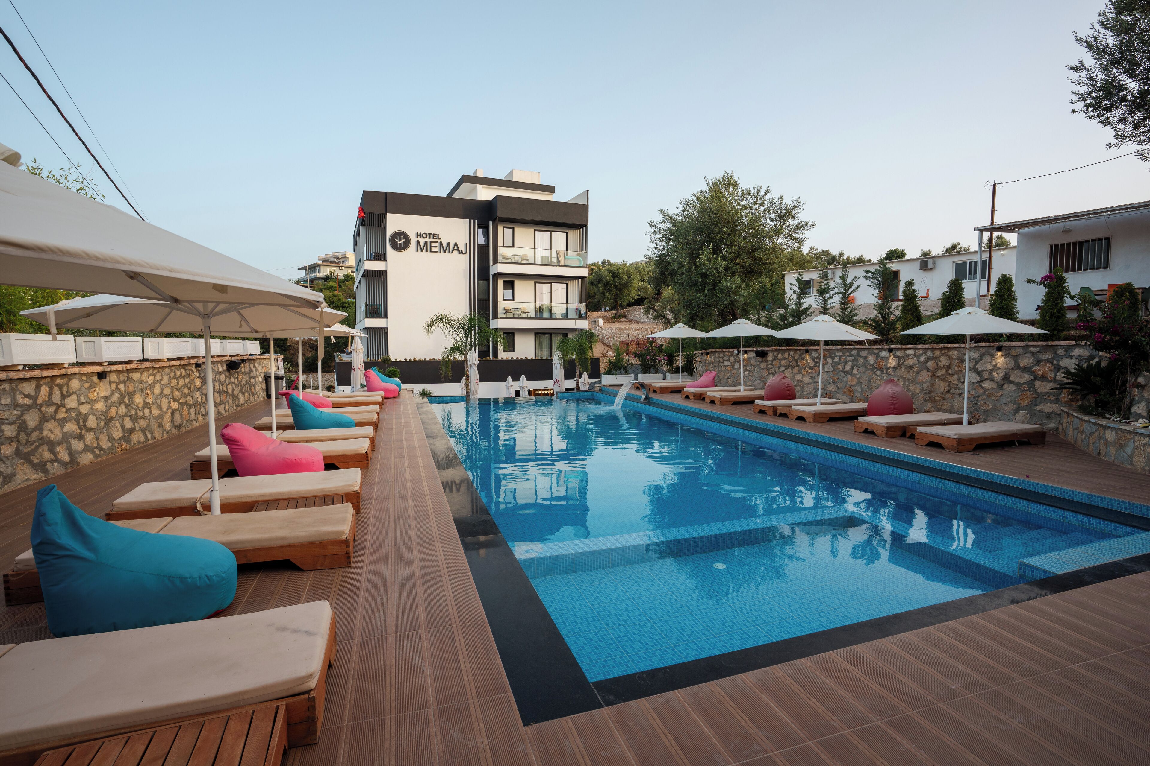 A hotel pool area with lounge chairs and umbrellas, a stone wall, and a building with a sign reading "HOTEL MEMAJ".