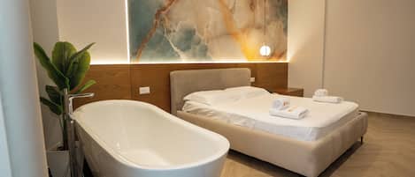 A modern bathroom with a freestanding bathtub, a bed with white linens, and a large abstract wall art piece.