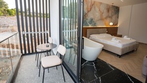Un dormitorio moderno con una cama grande, una bañera y un balcón acristalado con sillas.