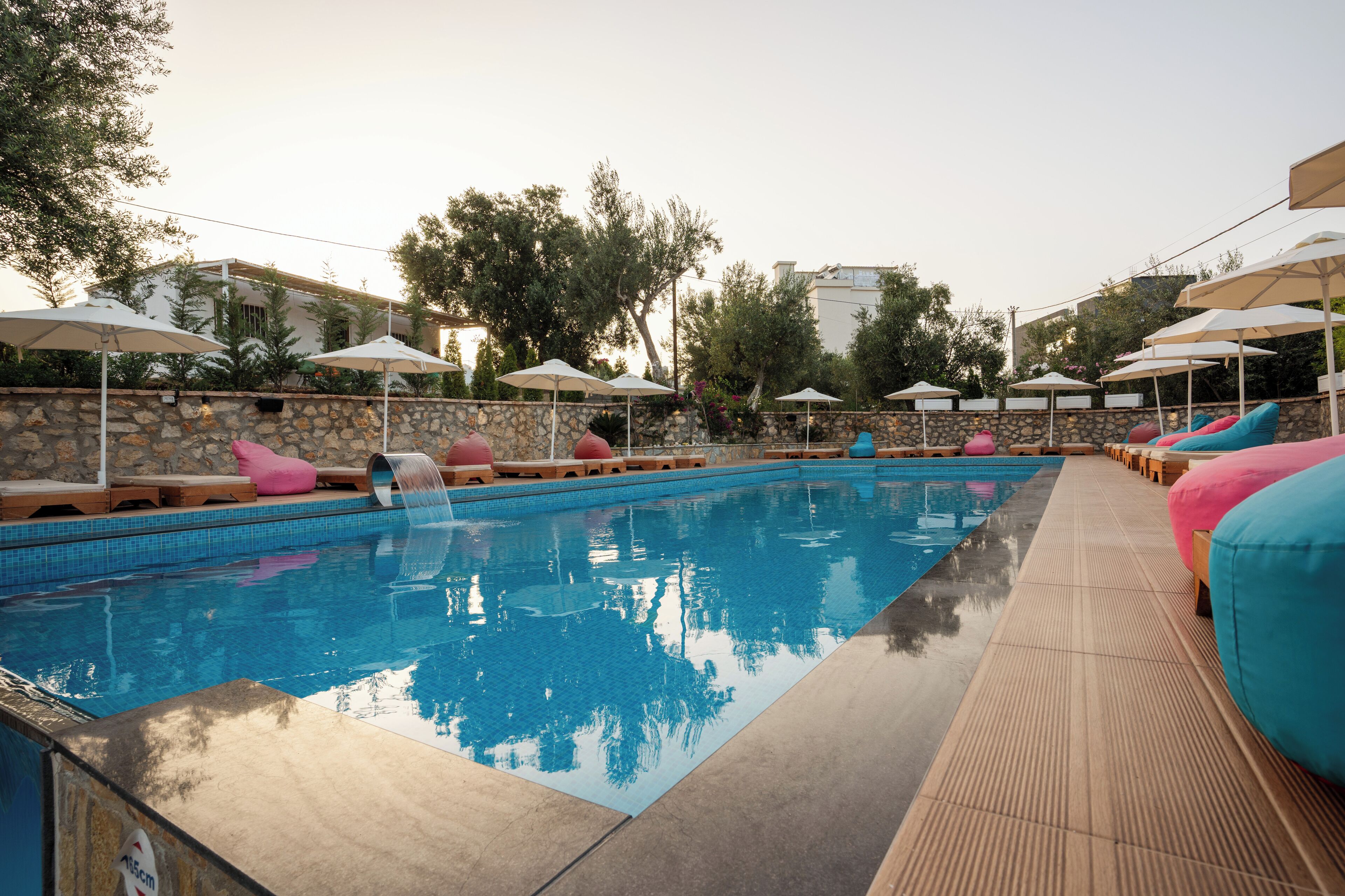 Piscina con sillas de descanso y sombrillas, rodeada de muros de piedra y árboles.