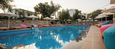 Piscina con sillas de descanso y sombrillas, rodeada de muros de piedra y árboles.