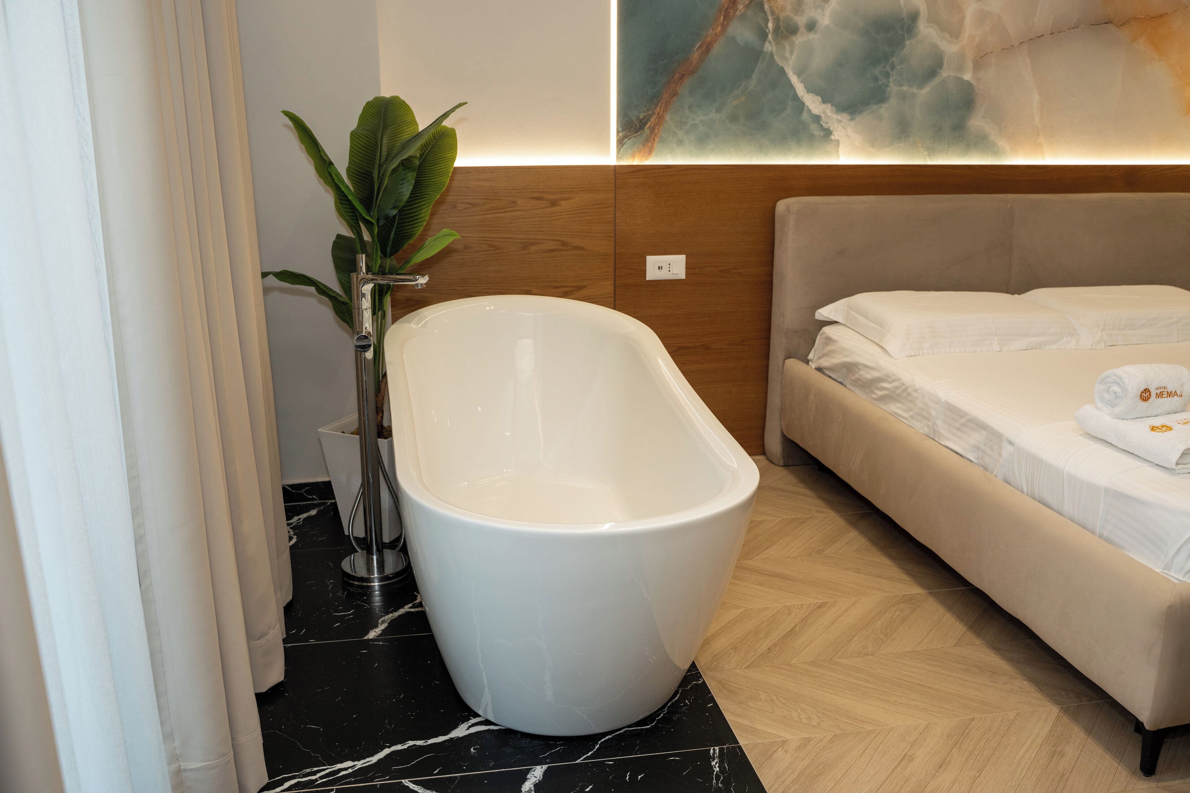 A modern bathroom with a freestanding bathtub, a potted plant, and a bed with a white headboard.