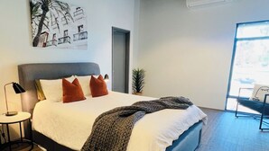 A modern bedroom with a large bed, a nightstand, a lamp, a potted plant, and a window with a view. -  Modern New Yorker Inspired Townhouse~ Full kitchen-Fast Wifi- Near Bunbury (Australind)