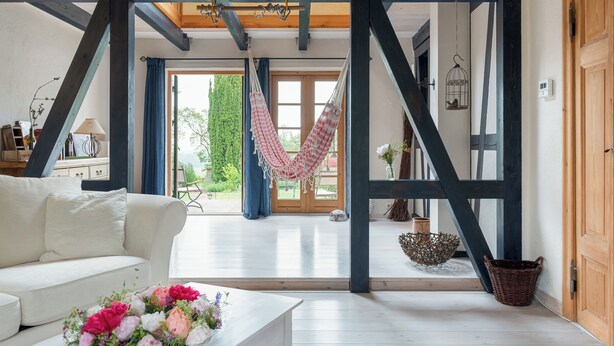 Ein geräumiges Wohnzimmer mit einem weißen Sofa, einem Couchtisch mit Blumen, einer Hängematte und Blick ins Freie.