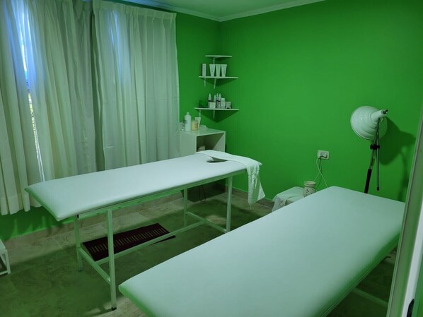 A medical examination room with two examination tables, a shelf with medical supplies, a white lamp, and a window with white curtains. - Intersur Independencia (Termas de Rio Hondo)