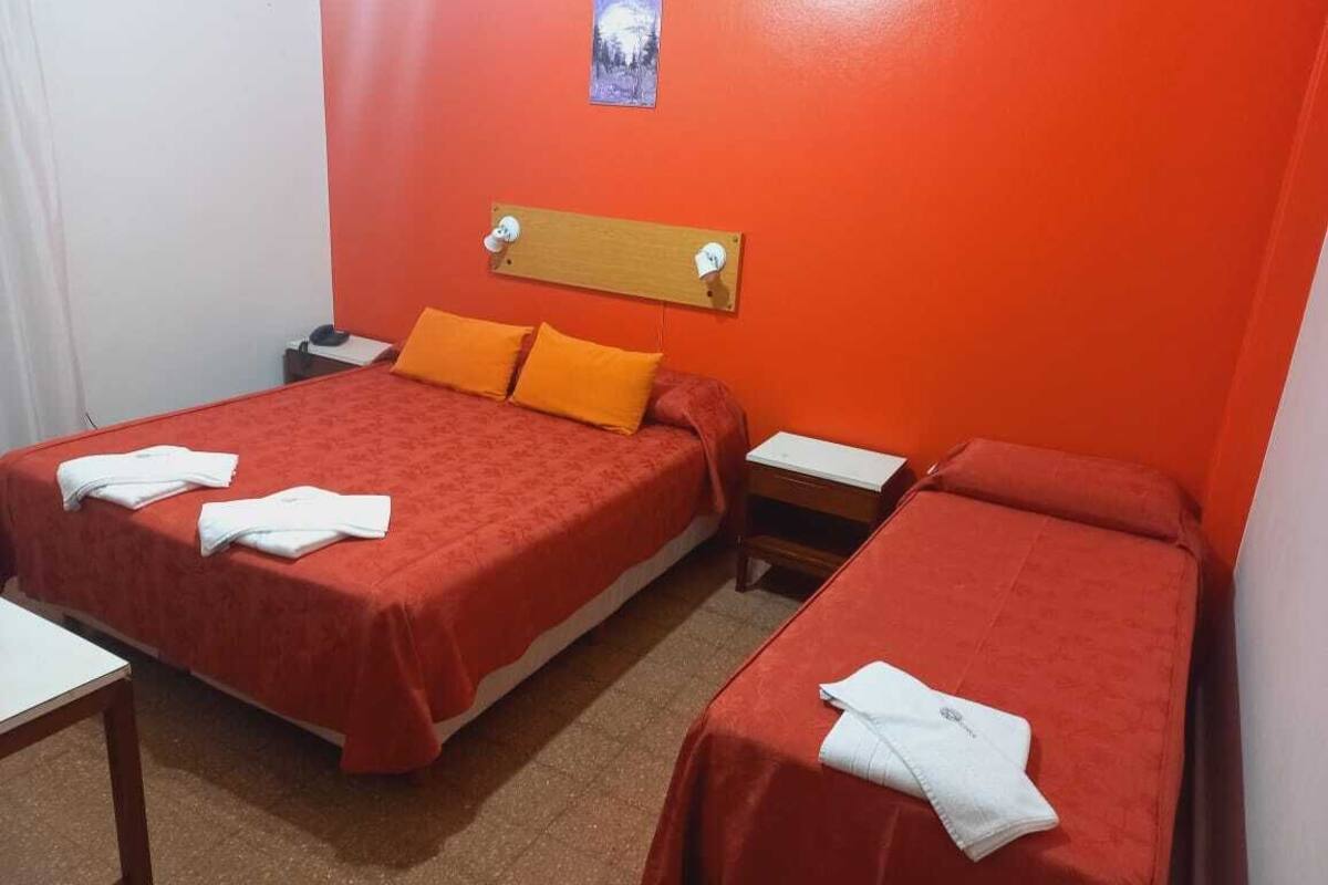 A room with two beds, each with a red bedspread and white towels. There is a wooden headboard above the beds and a small table between them.