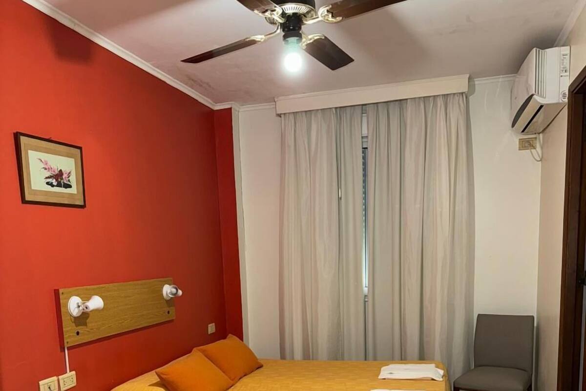 A hotel room with a bed, a desk, a chair, a ceiling fan, and an air conditioning unit.