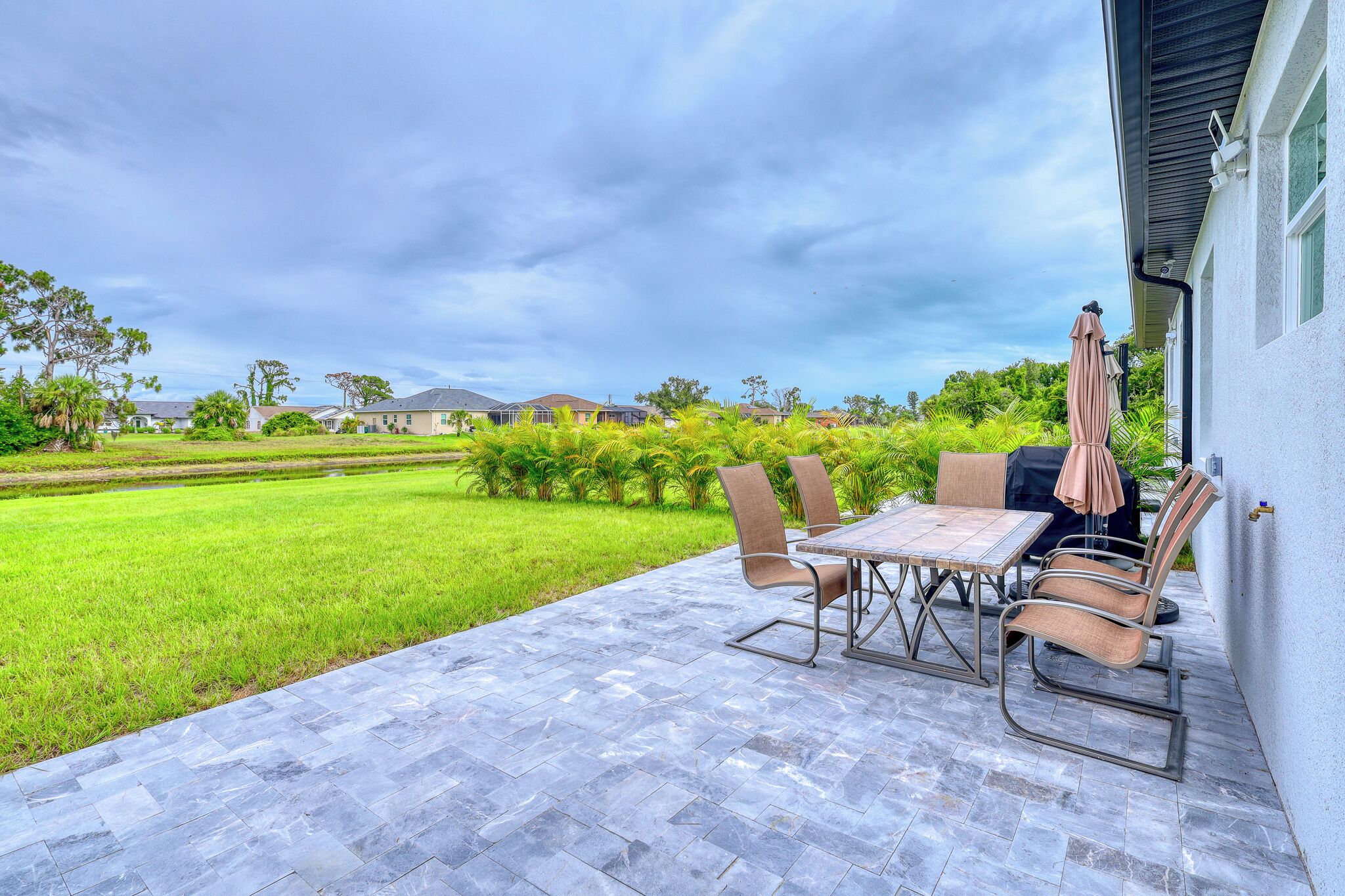 An outdoor patio with a table and chairs, a covered umbrella, and a view of a green lawn and houses in the background.