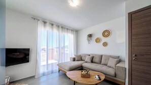 Ein modernes Wohnzimmer mit einer Eckcouch, einem Flachbildfernseher, einem Holztisch und einem großen Fenster mit durchsichtigen Vorhängen.