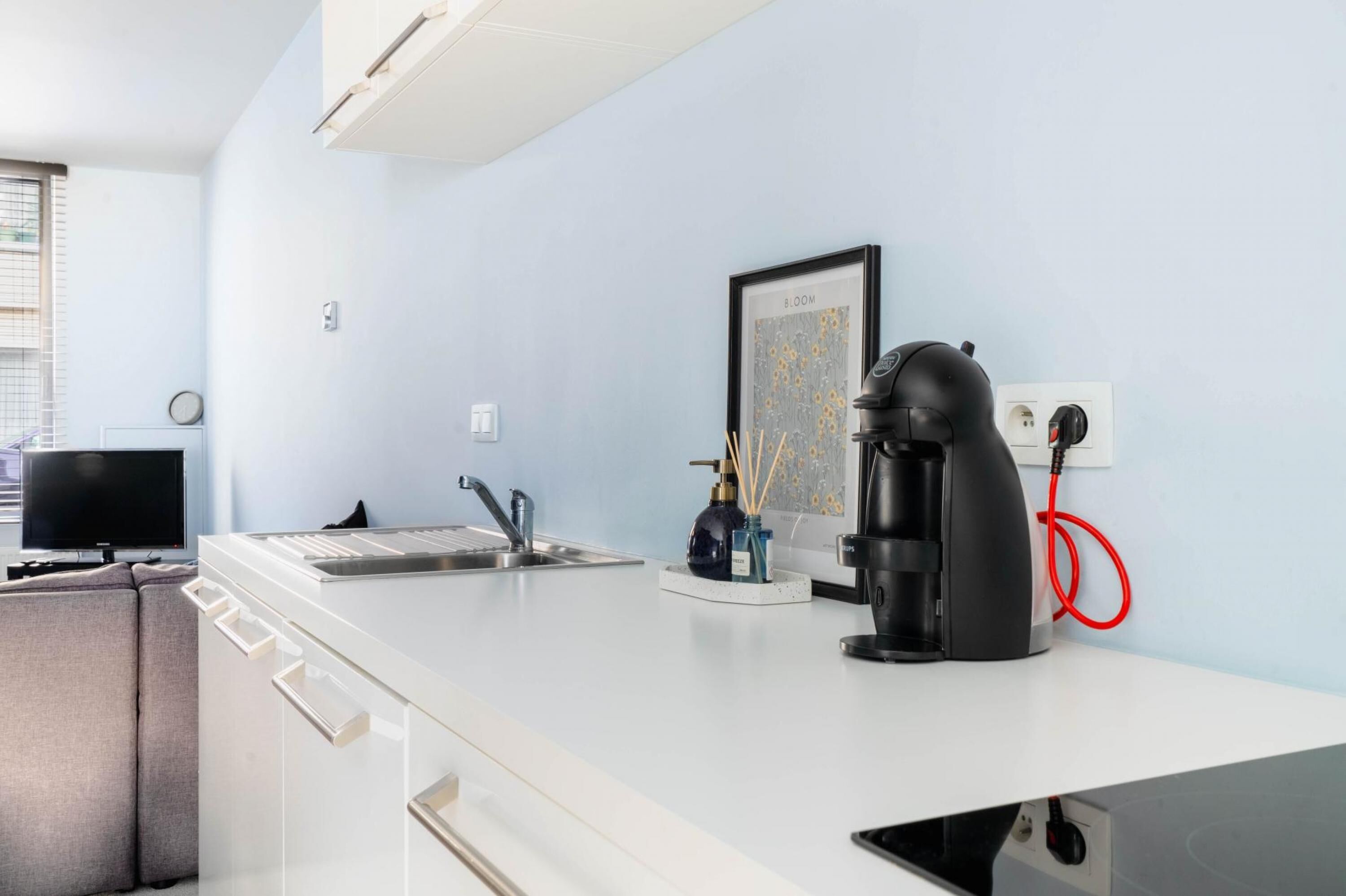Une cuisine moderne avec un plan de travail blanc, une machine à café, un évier et un tableau encadré au mur.