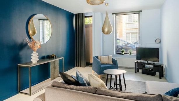 A modern living room with a blue accent wall, a round mirror, a sofa, a coffee table, a TV, and a console table with decorative items. - Charming Ostend Home Near Beach City Center (Ostend)