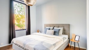 A bedroom with a large bed, a window with curtains, a bedside table with a lamp, and a wall-mounted light fixture. - Charming Ostend Home Near Beach City Center (Ostend)