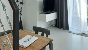 Un soggiorno moderno con un tavolo da pranzo in legno, un televisore a schermo piatto montato su una parete bianca, un'ampia finestra con tende leggere e una scala con ringhiere in metallo.