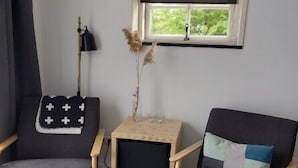 Ett rum med två grå fåtöljer, ett litet träbord med en vas och glas, och ett fönster med vit karm och persienner.