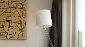 Un salon moderne avec un canapé en cuir noir, une lampe sur pied, un grand tableau abstrait et une vue par une fenêtre avec des rideaux.