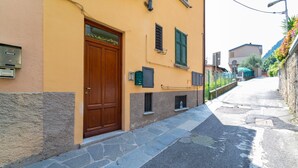 A narrow street with a yellow building featuring a wooden door and shutters, stone steps, and a metal mailbox. - Greenway Apartment by Wonderful Italy (Colonno)