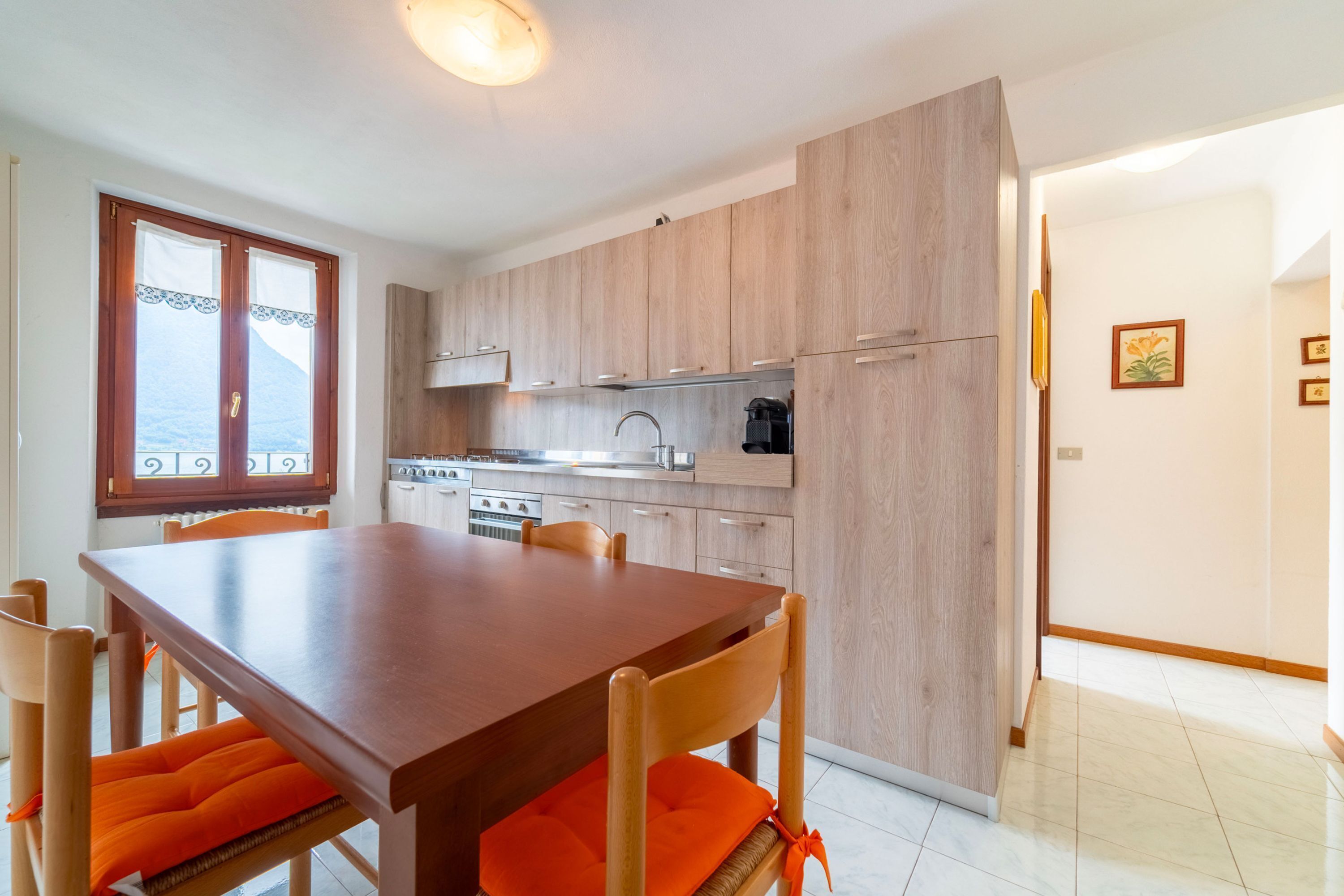 A modern kitchen with wooden cabinets, a dining table with orange cushions, and a view of the sea through a window.