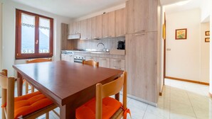 A modern kitchen with wooden cabinets, a dining table with orange cushions, and a view of the sea through a window. - Greenway Apartment by Wonderful Italy (Colonno)