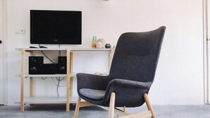 Une chambre moderne avec un téléviseur à écran plat fixé à un mur blanc, un fauteuil gris à revêtement en tissu et à pieds en bois, et une petite table agrémentée d’objets décoratifs.