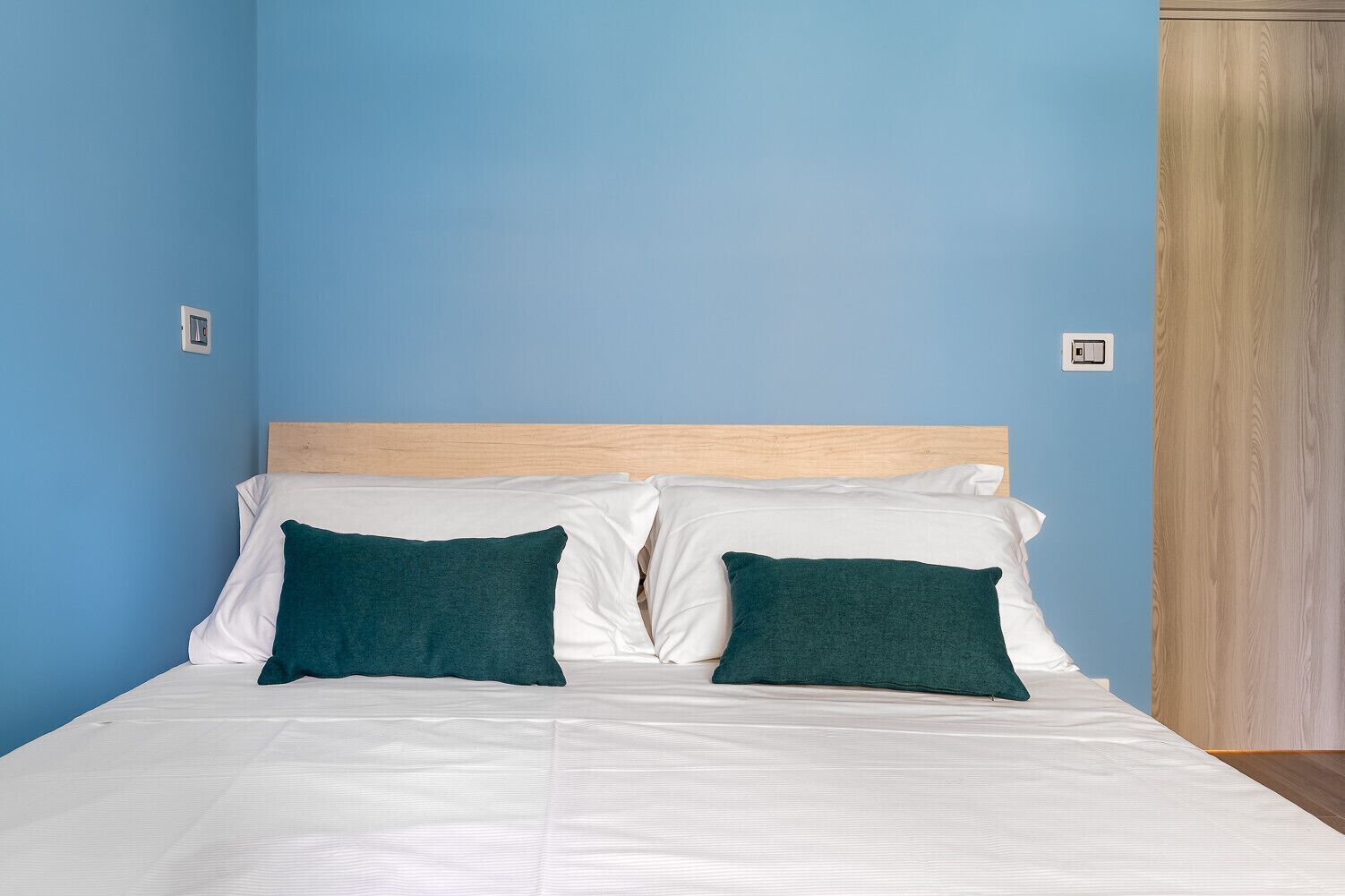 Una cama con ropa blanca y dos almohadas verde oscuro, un cabecero de madera y una pared azul.