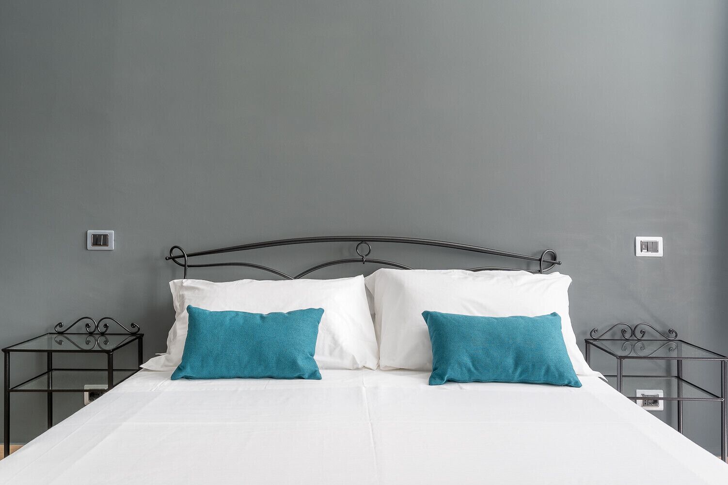 Una cama con ropa blanca y dos almohadas color turquesa, un cabecero de metal y dos mesitas de noche con superficie de vidrio.