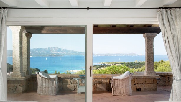 Een balkon met rieten stoelen, een stenen tafel en uitzicht op zee en bergen.