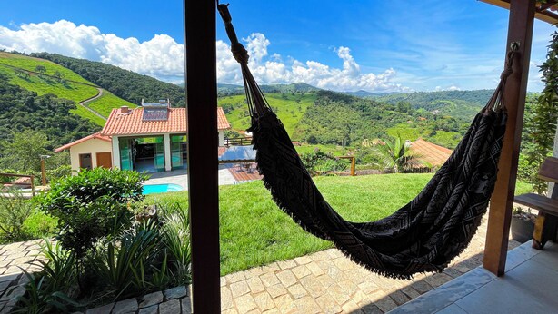 A hammock hanging under a wooden pergola with a view of a house and lush green hills in the background.