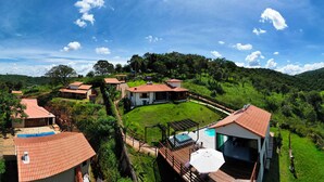A hillside property with multiple buildings, including a main house with a large deck and outdoor seating area, surrounded by lush greenery and hills.