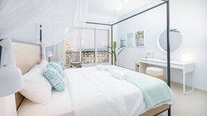 Un dormitorio moderno con cama con dosel, un balcón con vistas, un espejo redondo y un escritorio pequeño con lámpara.