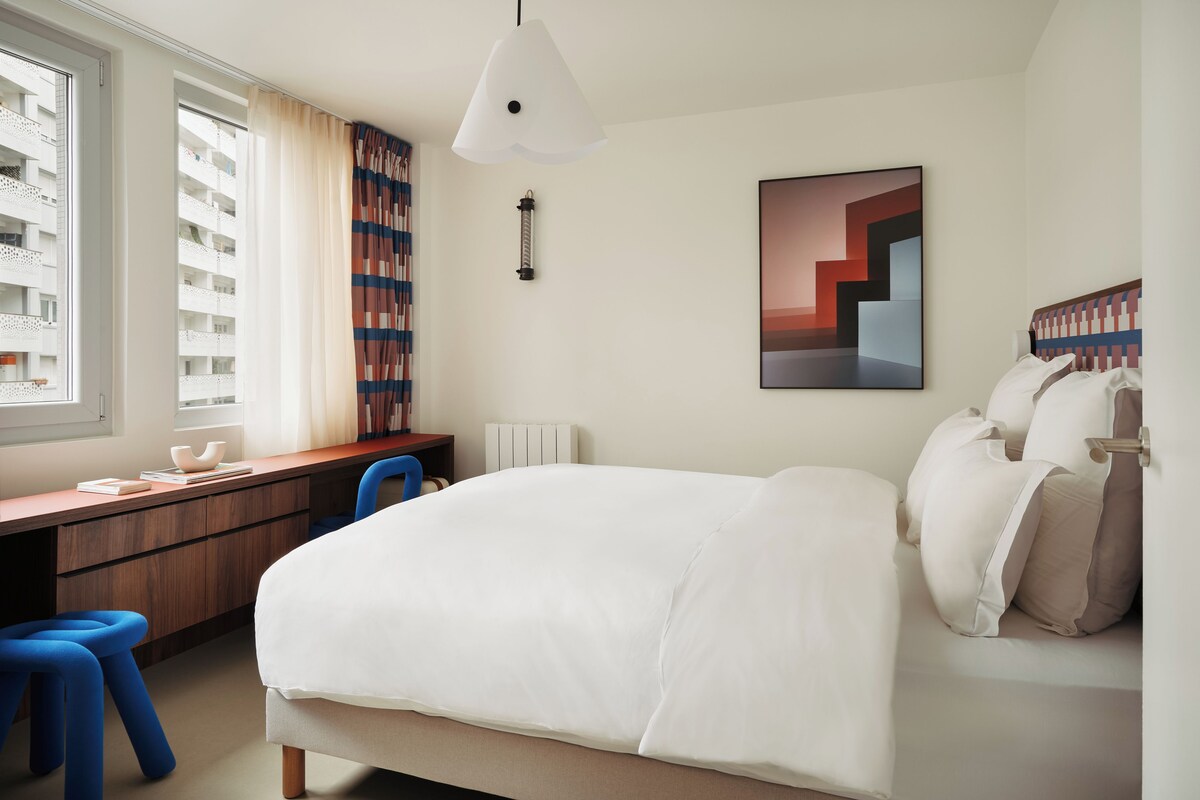 A modern hotel room with a large bed, a desk with a chair, a bookshelf, and a window with curtains.